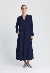 Marineblaues langärmeliges Kleid mit V-Ausschnitt, gestuftem Design, gerafften Details an den Ärmeln und seitlichen Taschen. Kombiniert mit metallicfarbenen flachen Schuhen.