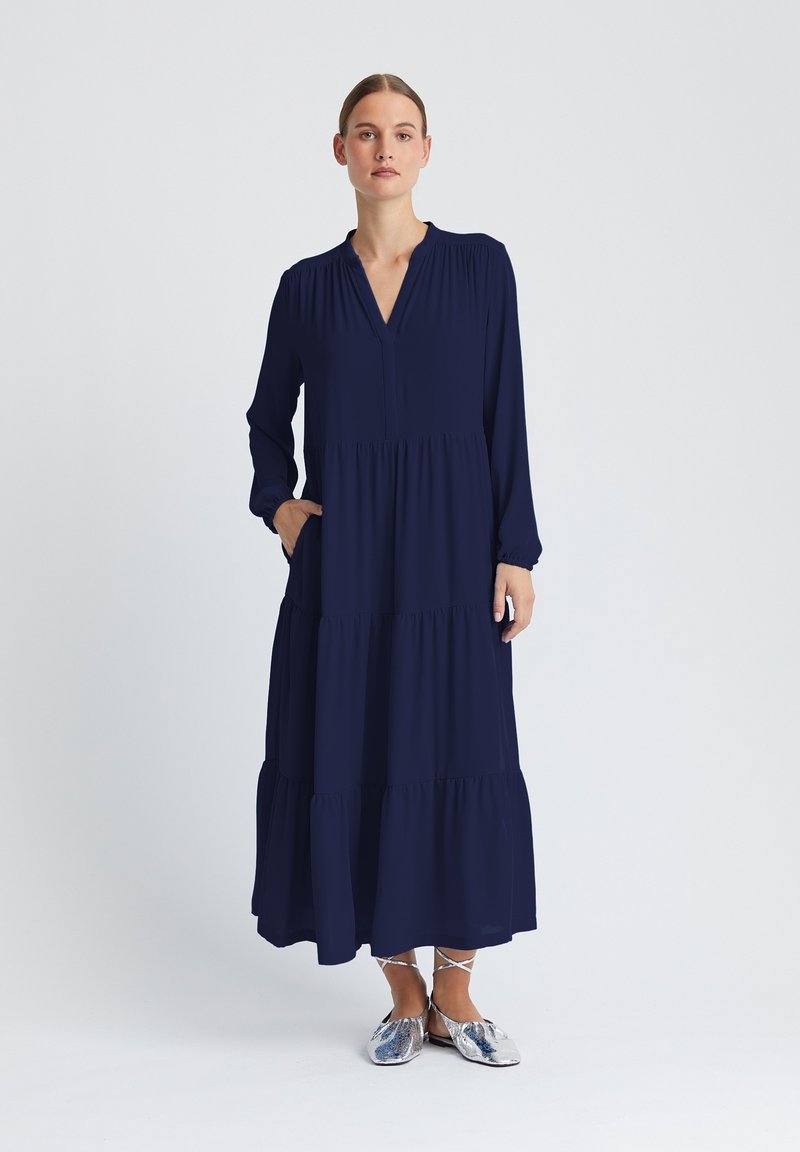 Marineblaues langärmeliges Kleid mit V-Ausschnitt, gestuftem Design, gerafften Details an den Ärmeln und seitlichen Taschen. Kombiniert mit metallicfarbenen flachen Schuhen.