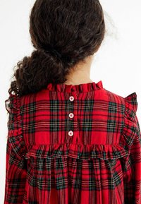 Vestido de cuadros rojos con bordes rizados, que presenta detalles de botones blancos en la parte posterior. La tela suave muestra un patrón a cuadros clásico en verde y negro.