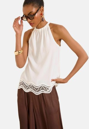 Vrouw die haar zonnebril afstelt, draagt een witte mouwloze top met kanten zoom, bruine wijde broekspijpen, een gouden armband en kleurrijke oorbellen.