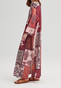 Long patchwork kimono with floral and paisley patterns in red, pink, and brown hues. Flared sleeves and a relaxed fit. Worn with brown sandals.