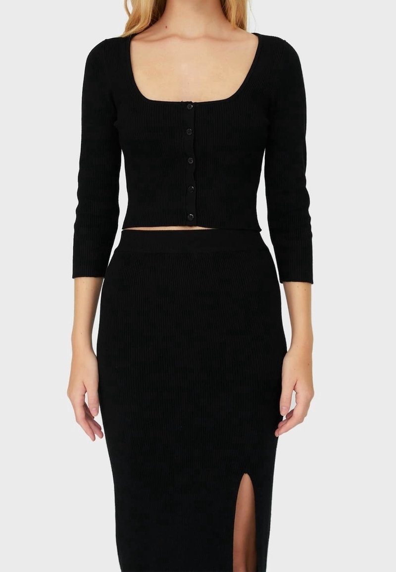 Femme portant un crop top côtelé noir à boutons avec des manches trois-quarts et une jupe assortie taille haute avec une fente à l'avant.