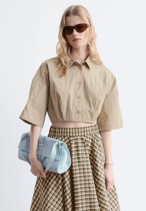 Femme portant des lunettes de soleil marron, une chemise courte beige à boutons, une jupe à carreaux, tenant un sac à main bleu clair sur un fond uni.