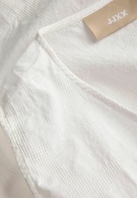 Λευκό ύφασμα με διακριτικά ανάγλυφα λουλουδένια και γεωμετρικά σχέδια, με ετικέτα ρούχου σε μπεζ χρώμα με το κείμενο «JUX».