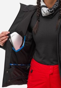 Intérieur de veste noire avec une poche bleue tenant des lunettes de ski blanches et argentées. Pantalons rouges avec fermeture à bouton et texture en tissu noir.