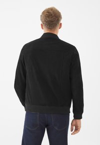 Svart mocka bomberjacka med ribbad krage och ärmslut, med en mjuk textur och minimalistisk design. Bärs över blå denimjeans.