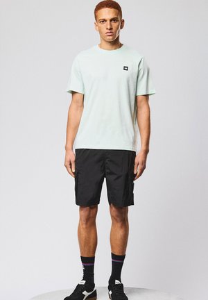 Junger Mann, der vor einem schlichten hellen Hintergrund steht und ein hellblaues T-Shirt, schwarze Cargo-Shorts, schwarze Socken und schwarze Sneakers trägt.