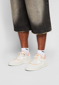 Jambes portant un short en denim noir délavé, des chaussettes blanches Puma et des baskets blanches Puma avec des accents pêche sur un fond gris clair.