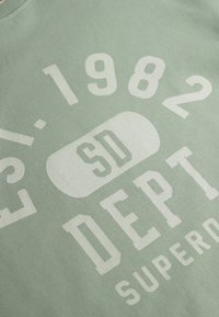 Sudadera verde menta con un gran texto gráfico blanco, que incluye "EST. 1982", "SD" y "DEPT." en diferentes tipografías. Textura suave y lisa.