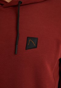 Sudadera roja hecha de tejido suave, que presenta un parche cuadrado negro con un diseño de línea naranja y cordones negros.
