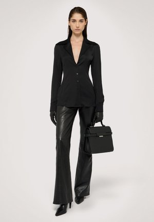 Czarna bluzka z długim rękawem i głębokim dekoltem w kształcie V, zestawiona z dopasowanymi czarnymi skórzanymi spodniami i eleganckimi czarnymi szpilkami, trzymająca usztywnioną czarną torebkę.