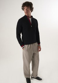 Felpa nera con zip, dal design testurizzato e dettagli rossi al colletto, abbinata a pantaloni beige chiaro a taglio morbido e scarpe nere.