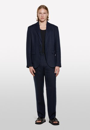 Costume marin composé d'un blazer à un bouton et d'un pantalon droit en tissu léger, associé à un débardeur noir et des sandales.