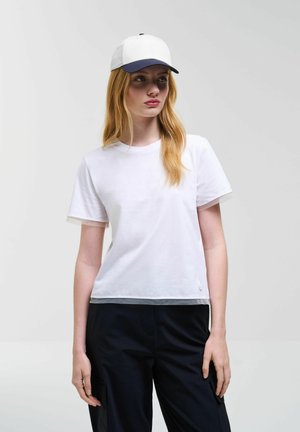 Giovane donna con capelli lunghi che indossa una t-shirt bianca, pantaloni neri e un berretto da baseball bianco con visiera blu scuro, in piedi davanti a uno sfondo semplice.