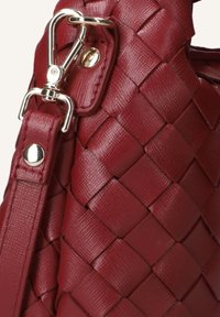 Detail van een rode geweven leren handtas met een metalen clip en band, voorzien van gestructureerde oppervlakken en gepolijste accenten.