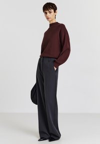 Maglione in maglia color bordeaux con collo alto, abbinato a pantaloni a gamba larga grigio scuro. Scarpe nere con punta a punta. Borsa nera strutturata tenuta sotto il braccio.