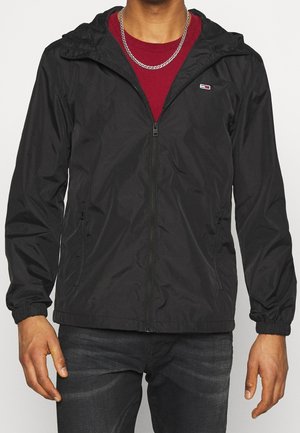 Homme portant une veste à capuche noire zippée sur une chemise rouge avec un collier chaîne argentée, jean foncé, debout devant un fond uni.