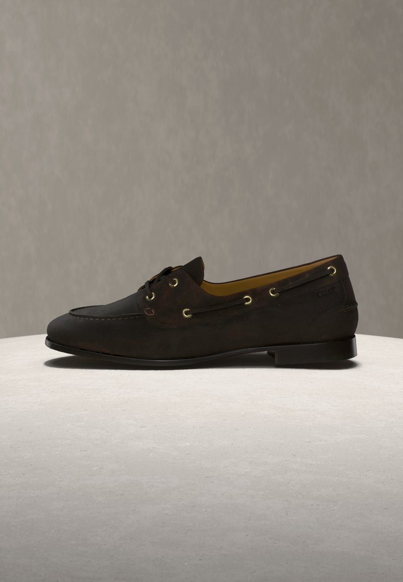 Σκούρο καφέ δερμάτινο μοκασίνι με λεπτομέρειες από κορδόνια και χρυσά τρύπες, τοποθετημένο πάνω σε φωτεινή πέτρινη επιφάνεια με απλό μπεζ φόντο.