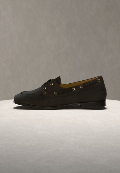 Σκούρο καφέ δερμάτινο μοκασίνι με λεπτομέρειες από κορδόνια και χρυσά τρύπες, τοποθετημένο πάνω σε φωτεινή πέτρινη επιφάνεια με απλό μπεζ φόντο.