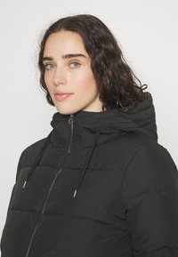 Chaqueta acolchada negra con cuello alto, cierre de cremallera, capucha ajustable y tela texturizada, que presenta detalles de costura horizontal.