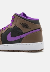 Bruna och svarta Air Jordan mid-top sneakers med lila snörning, lila Nike swoosh och vit sula på en vit bakgrund.