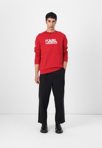 Sudadera roja con "KARL LAGERFELD" impresa en blanco, combinada con pantalones de pierna ancha negros y zapatos negros, sobre un fondo liso.