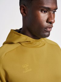 Zbliżenie mężczyzny w musztardowożółtej bluzie z kapturem z logo „hummel” na piersi, patrzącego w bok.
