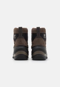 Botas impermeables de ante marrón y tela negra con suelas de goma, que cuentan con lengüetas y un logo en la parte trasera. Diseño corto y redondeado.