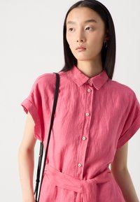 Camicia di lino rosa con colletto, maniche corte e una cintura con legatura in vita. Dettagli notevoli includono la chiusura con bottoni e un tessuto testurizzato.