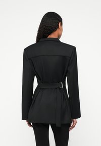 Mustas kohandatud bleiser, millel on vööga talje, krae ja pikad varrukad. Omab siledat tekstuuri ja struktureeritud disaini, kohandatud sobivust.