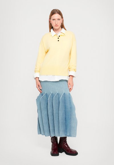 Maglione giallo a maglia con tre bottoni neri, indossato sopra una camicia bianca. Gonna in denim plissettata di un blu chiaro con bordi sfilacciati. Stivali alla caviglia bordeaux.