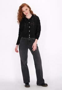 Cardigan nero a maglia con chiusura a bottone e vestibilità ampia, abbinato a jeans grigi a gamba dritta e stivali neri. Sfondo neutro.