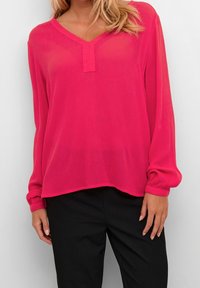 Femme portant un chemisier à manches longues de couleur rose vif avec un décolleté en V et un pantalon noir, se tenant devant un fond clair uni.