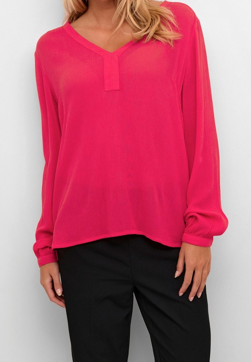 Femme portant un chemisier à manches longues de couleur rose vif avec un décolleté en V et un pantalon noir, se tenant devant un fond clair uni.