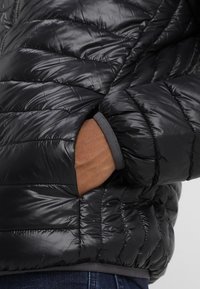 Svart puffjacka med quiltad textur, sidofickor och elastiska ärmslut. Materialet är glänsande och slätt med en figursydd design.