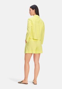 Yellow linen jacket and matching shorts, featuring a relaxed fit, double-stitched hems, and side pockets. Worn with patterned slip-on sandals.