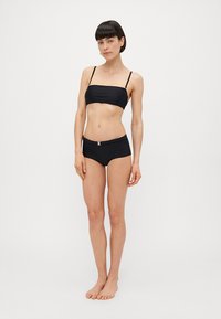 Melnā bikini komplektā ietilpst bezplecu tops un augsta jostasvietas apakšdaļa ar sudraba sprādzi. Materiāls ir gluds un elastīgs.