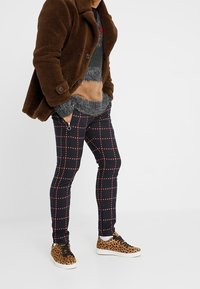 Bruine fleece jas, grijs gemusterd Sweater, marineblauwe geruite broek met rode lijnen, en sneakers met luipaardprint, zwarte veters en witte zolen.