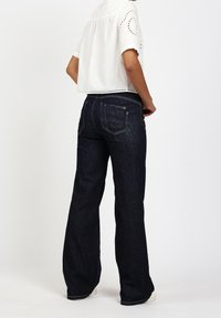 Jeans de mezclilla oscuros acampanados con costuras en contraste y una cintura de altura media, combinados con una blusa corta blanca con detalles de ojal.