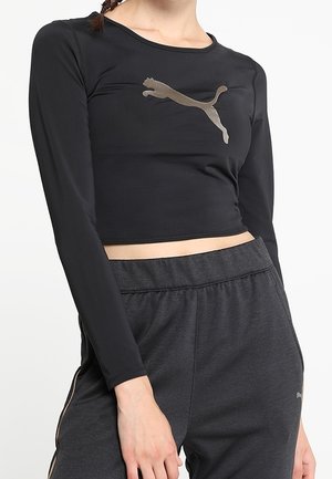 Femme portant un crop top noir à manches longues avec un logo Puma argenté et un pantalon taille haute gris foncé avec des poches.