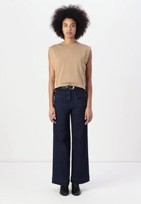 Top sin mangas beige combinado con jeans de pierna ancha azul oscuro, que cuentan con bolsillos frontales y un cinturón negro. La modelo lleva botas negras hasta el tobillo.