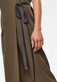 Vestido verde oliva con un bolsillo lateral, tejido texturizado y un cinturón trenzado negro y gris atado a la cintura.