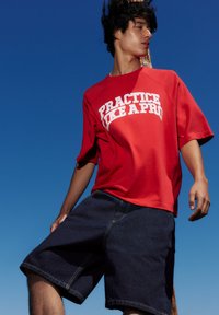 Jeune homme en chemise rouge avec le texte "PRACTICE LIKE A PRO" et un short en jean foncé se tient devant un ciel bleu clair.