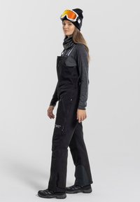 Black snow overalls with adjustable straps, side pockets, and reinforced knee panels. Paired with a dark gray long-sleeve top and black beanie.