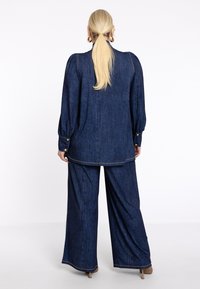Damenoutfit aus Denim mit einer locker sitzenden Bluse mit Puffärmeln und weitem Hosenbeinen. Das Material ist dunkelblau mit kontrastierenden Nähten und goldenen Knöpfen.