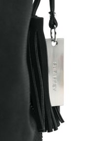 Juoda odinė kutas ir metalinė žymė su įspaustu užrašu „SURI FREY“, pritvirtinta prie juodos tekstūruotos paviršiaus.