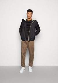 Giacca nera con cappuccio e foderata in pile crema, con chiusura a zip, abbinata a un maglione a strisce scure, pantaloni beige e sneaker bianche.