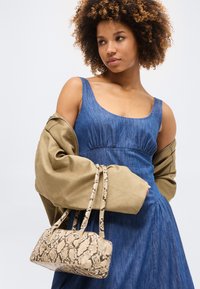 Γυναίκα με σγουρά μαλλιά φοράει μπλε φόρεμα χωρίς μανίκια και καφέ μπουφάν πέριξ των ώμων, κρατώντας μια μπεζ τσάντα με σχέδιο φολιδωτού δέρματος φιδιού.