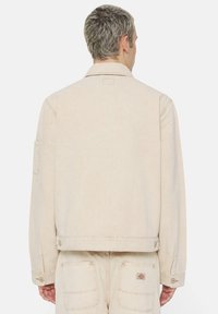 Giacca di jeans beige chiaro con colletto classico, maniche lunghe e chiusura con bottoni. Presenta un yoke posteriore e dettagli con tasche a toppa.