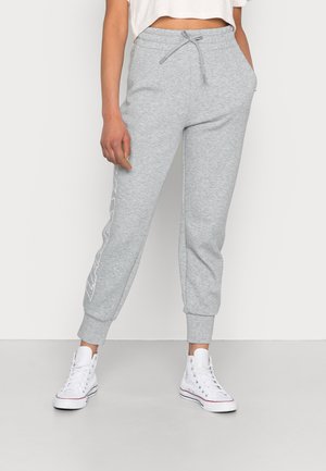 Kvinde iført lysegrå joggebukser med tekst i siden og hvide høje sneakers, stående mod en ensfarvet lys baggrund.